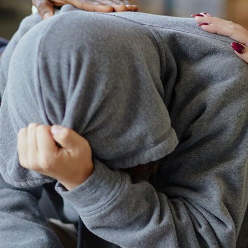 A stressed out teenage boy with his hood up and face in palms, a gentle reassuring hand on his back